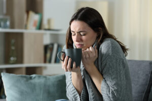 Woman sitting on a couch holding a mug, frowning in discomfort as she tries to hold her sweater around herself to keep warm because the furnace isn't keeping the house comfortable in winter.